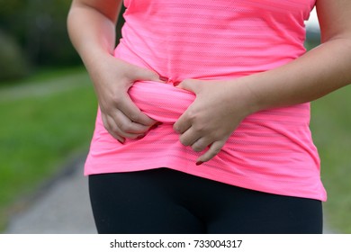 Woman Wearing A Pink Shirt Outdoors Holding A Roll Of Excess Fat Around Her Stomach Area Pinched In Her Hands In A Close Up Torso View