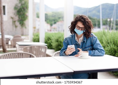 Woman Wearing Protective Mask In Empty Cafe Outdoors. Girl Using Mobile Phone. Social Distancing During Covid 19 Quarantine. Lifestyle Moment. Restaurant Terrace With Coronavirus Lockdown Restrictions