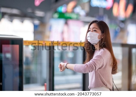 Woman wearing protective mask looks at watch on her wrist at train station platform, waiting someone who is in late, mass rapid transit and crowd at the station in the background