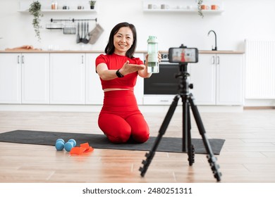 Woman wearing red fitness outfit kneeling on black mat filming online workout tutorial in bright kitchen setting. She holds large water bottle, smiling. Camera on tripod captures entire scene. - Powered by Shutterstock