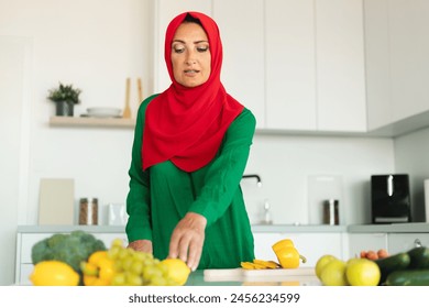 A woman wearing a red and green hijab is shown in the act of chopping a variety of vegetables on a cutting board. - Powered by Shutterstock