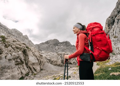 A woman wearing a red jacket and headband stands on a rocky trail, holding trekking poles, surrounded by mountains and overcast skies. - Powered by Shutterstock