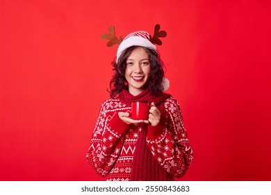 The woman is wearing a red sweater with a white pattern and a red scarf. She has a red mug in her hands and is looking at the camera with a smile on her face. The background is a solid red color. - Powered by Shutterstock