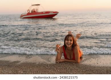 A woman wearing an red swimsuit is lying on the sandy shore, holding a glass of champagne, with a smile on her face. In the background, a red speedboat is seen floating on the tranquil sea - Powered by Shutterstock