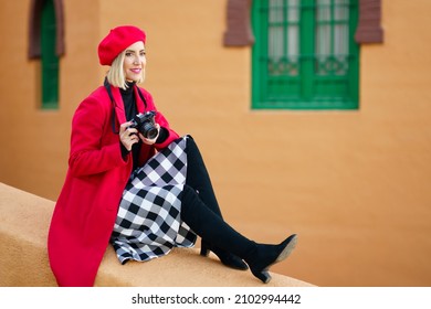 Woman wearing red winter clothes, taking pictures with an SLR camera sitting on a city wall. - Powered by Shutterstock