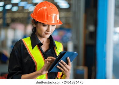 A woman wearing a safety vest and an orange helmet is looking at a tablet. She is likely a worker in a factory or warehouse, using the tablet to check information or instructions - Powered by Shutterstock