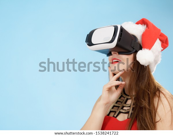 Woman wearing santa claus hat exploring space with
virtual reality goggles headset. Amazed girl watching 3d film tour
in vr glasses box. New generation cyber christmas concept, on
blue.
