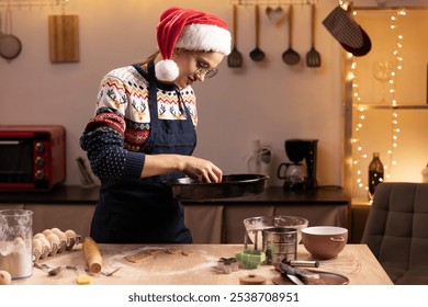 Woman wearing Santa hat and festive sweater carefully placing raw gingerbread cookies on baking tray for Christmas. Baking tools, dough, and cookie cutters spread across kitchen table - Powered by Shutterstock