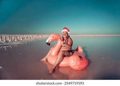 A woman wearing a Santa hat sits on a pink flamingo float in a body of water. He is smiling. There are white posts in the background.