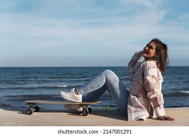 Woman wearing shirt and jeans sitting on the boardwalk next to her longboard. Young girl sitting sideways leaning on her skateboard with the sun on her back and fresh air.