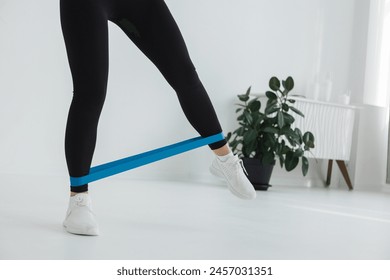 The woman, wearing sportswear made of jersey fabric, is performing exercises on the floor with a blue resistance band wrapped around her thigh and knee - Powered by Shutterstock