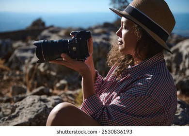 A woman, wearing a straw hat and plaid shirt, sits outdoors holding a camera. She is enjoying nature while photographing the rocky landscape. A sense of adventure and exploration. - Powered by Shutterstock