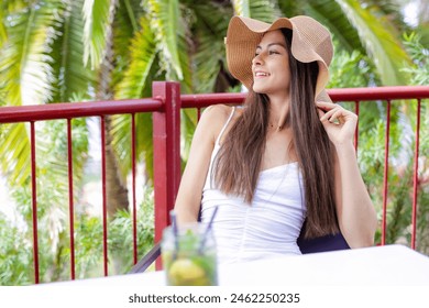 Woman wearing straw hat is sitting at table with drink in front of her. Thoughtful woman thinking and looking at side sitting in bar or home terrace - Powered by Shutterstock