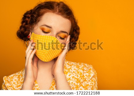 Woman wearing stylish handmade protective face mask posing on orange background. Monochrome color outfit. Model with bold eyes makeup. Fashion during quarantine of coronavirus. Copy space for text