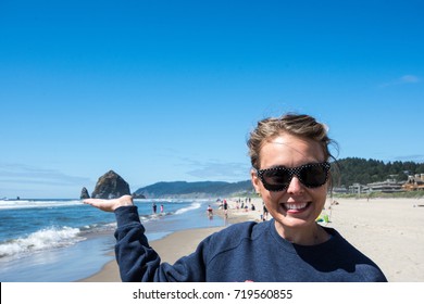 Woman Wearing Sunglasses Pretends To Hold Up A Seastack Rock In Cannon Beach, Oregon On The Beach On A Sunny Day. Forced Perspective View