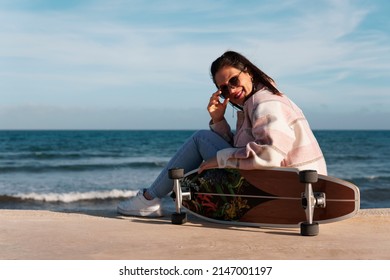 Woman wearing sunglasses, shirt and jeans sitting on the boardwalk next to her longboard. Young girl sitting sideways leaning on her skateboard with the sun on her back and fresh air.