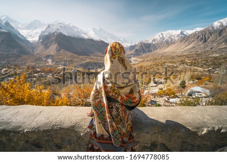 A woman wearing traditional dress sitting on wall and looking at Hunza valley in autumn season, Gilgit Baltistan in Pakistan, Asia