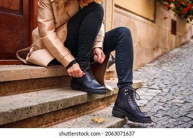 Woman wearing trench coat sitting on staircase and tying shoelace on her ankle boot. Autumn fashion collection. Trendy black leather shoe. Street style