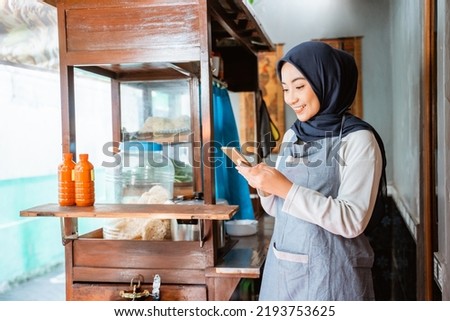 woman wearing a veil wearing an apron using a cellphone while selling at a chicken noodle cart