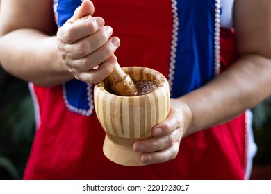 Woman wearing vintage red and blue apron is holding a wooden mortar and pestle in her hands while preparing a traditional Macedonian meal with roasted paprika and garlic.