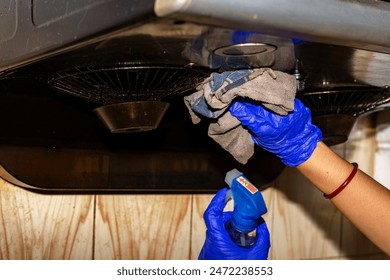 A woman wearing waterproof gloves uses a chemical spray to clean a kitchen chimney, demonstrating proper cleaning techniques and safety measures in a modern home kitchen. - Powered by Shutterstock