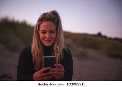 Woman Wearing Wetsuit Sitting On Beach Using Mobile Phone As Sun Sets Behind Her