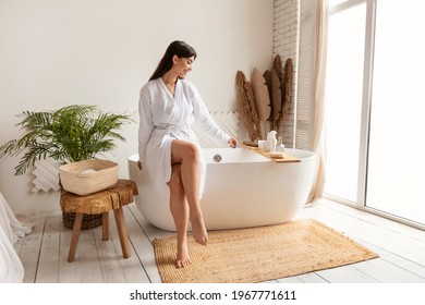 Woman Wearing White Bathrobe Preparing Bath Adding Sea Salt Sitting On Bathtub In Modern Bathroom Indoors. Spa Day And Female Beauty Routine Concept. Free Space For Text - Powered by Shutterstock