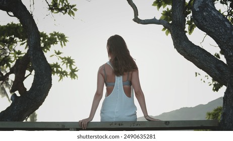 A woman wearing a white dress is seated on a wooden fence, her legs dangling in the air. She gazes into the distance, with a serene expression on her face. - Powered by Shutterstock