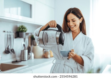 A woman wearing a white robe is pouring tea from an electric kettle in a modern kitchen, smiling happily. The scene suggests comfort, relaxation, and a peaceful morning routine. - Powered by Shutterstock