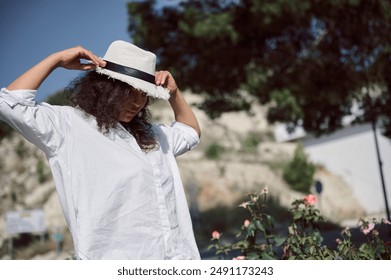 Woman wearing white shirt adjusting her hat outdoors on a sunny day. A relaxed and serene moment captured amidst nature. - Powered by Shutterstock