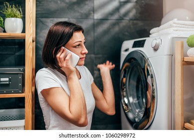 Woman Wearing White Shirt Sitting At Landry Room By The Washing Machine And Calling For Appliance Repair Service