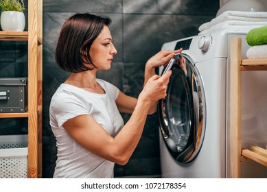 Woman Wearing White Shirt Using Smart Phone To Control Washing Machine At Laundry Room