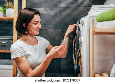 Woman Wearing White Shirt Using Smart Phone To Control Washing Machine At Laundry Room