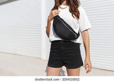 Woman, wearing white t-shirt, black shorts and fanny pack or waist pack, standing outdoor near white wall. Details of stylish trendy basic minimalistic casual outfit. Street fashion. No face.