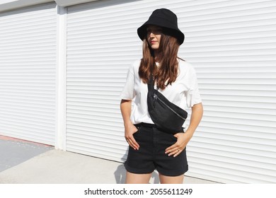 Woman, wearing white t-shirt, black shorts, fanny pack or waist pack and bucket hat, standing outdoor near white wall. Details of stylish trendy basic minimalistic casual outfit. Street fashion. - Powered by Shutterstock
