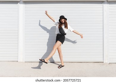 Woman, wearing white t-shirt, black shorts, fanny pack or waist pack, bucket hat and flat sandals, standing outdoor near white wall. Stylish trendy basic minimalistic casual outfit. Street fashion. - Shutterstock ID 2057725721