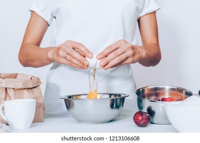 Woman wearing white t-shirt breaking raw egg while cooking pie. Female standing at table with metal bowl, fresh fruits, sack of flour, cup and preparing dough for plum cake. - Powered by Shutterstock