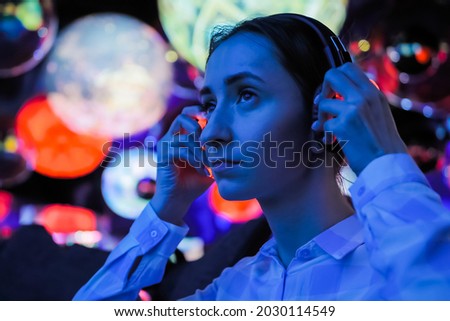 Woman wearing wireless black headphones and looking around in dark room of interactive exhibition or museum with colorful illumination. Futuristic, entertainment, immersive concept Stockfoto © 