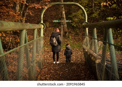 A Woman Wearing A Wooly Hat And Green Pack Back Stands Next To Her Daughter In Front Of A Sign Post With A Bridge On The Foreground And The Ground Covered In Brown Autumn Coloured Leaves In A Forest