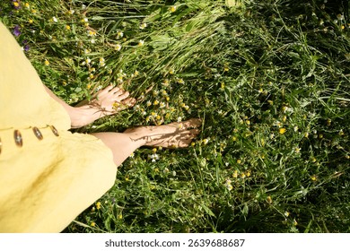 Woman wearing yellow dress standing barefoot on green grass with chamomile flowers enjoying summer