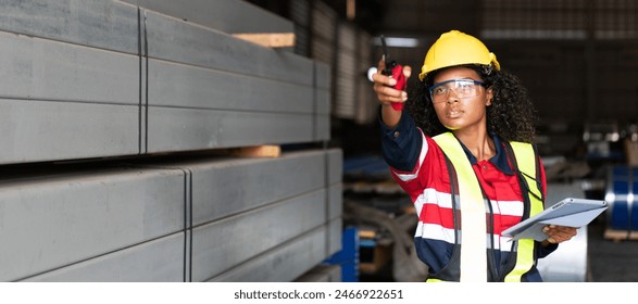 A woman wearing a yellow helmet and a red vest is pointing at something on a clipboard. She is wearing safety gear and is standing in a warehouse - Powered by Shutterstock