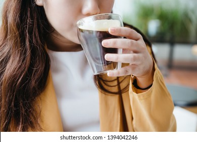 Woman Wearing Yellow Jacket Sipping Frothy Nitro Cold Brew Coffee In Drinking Glass With Green Blur Background.