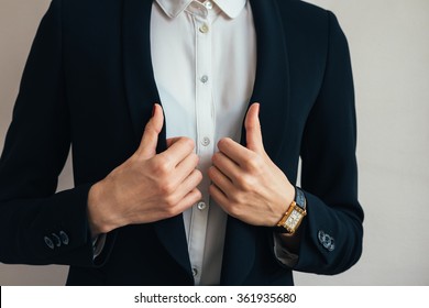 Woman Wears A Business Suit Jacket. In Her Hand Wristwatch. Close-up Of A Woman In A Business Suit And Wristwatch.