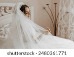 A woman in a wedding dress is sitting on a bed with a veil over her head. Concept of elegance and romance, as the bride is getting ready for her wedding day