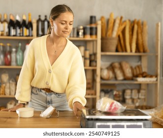 Woman weighs cheese in a grocery store against the background of shelves with food. Selection and purchase of different types of cheese made from natural milk - Powered by Shutterstock