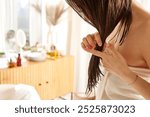 A woman with wet hair is inspecting the ends of her hair while standing in a bright, airy bathroom. She is wrapped in a towel, and there are beauty products on a wooden dresser in the background.