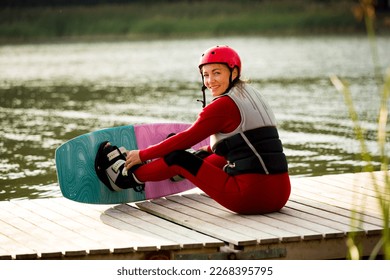 Woman in wetsuit, helmet and life vest sitting with wakeboard on a wooden pier. Sunny summer day. Safety in sport. Water sports in Finland. Insurance concept
