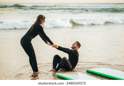 A woman in a wetsuit reaches out to help a man who has fallen off his surfboard in the shallows of a beach. He is sitting in the water with his surfboard nearby, while she is standing in the sand. - Powered by Shutterstock