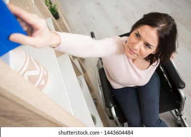 Woman In Wheelchair Reaching For Book From High Shelf