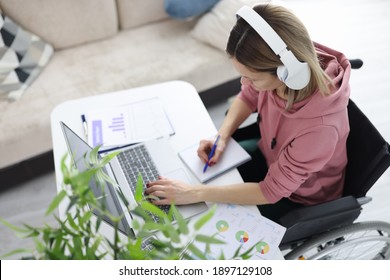 Woman in wheelchair sits at work desk wearing headphones and makes notes in notebook. Remote service for people with disabilities concept